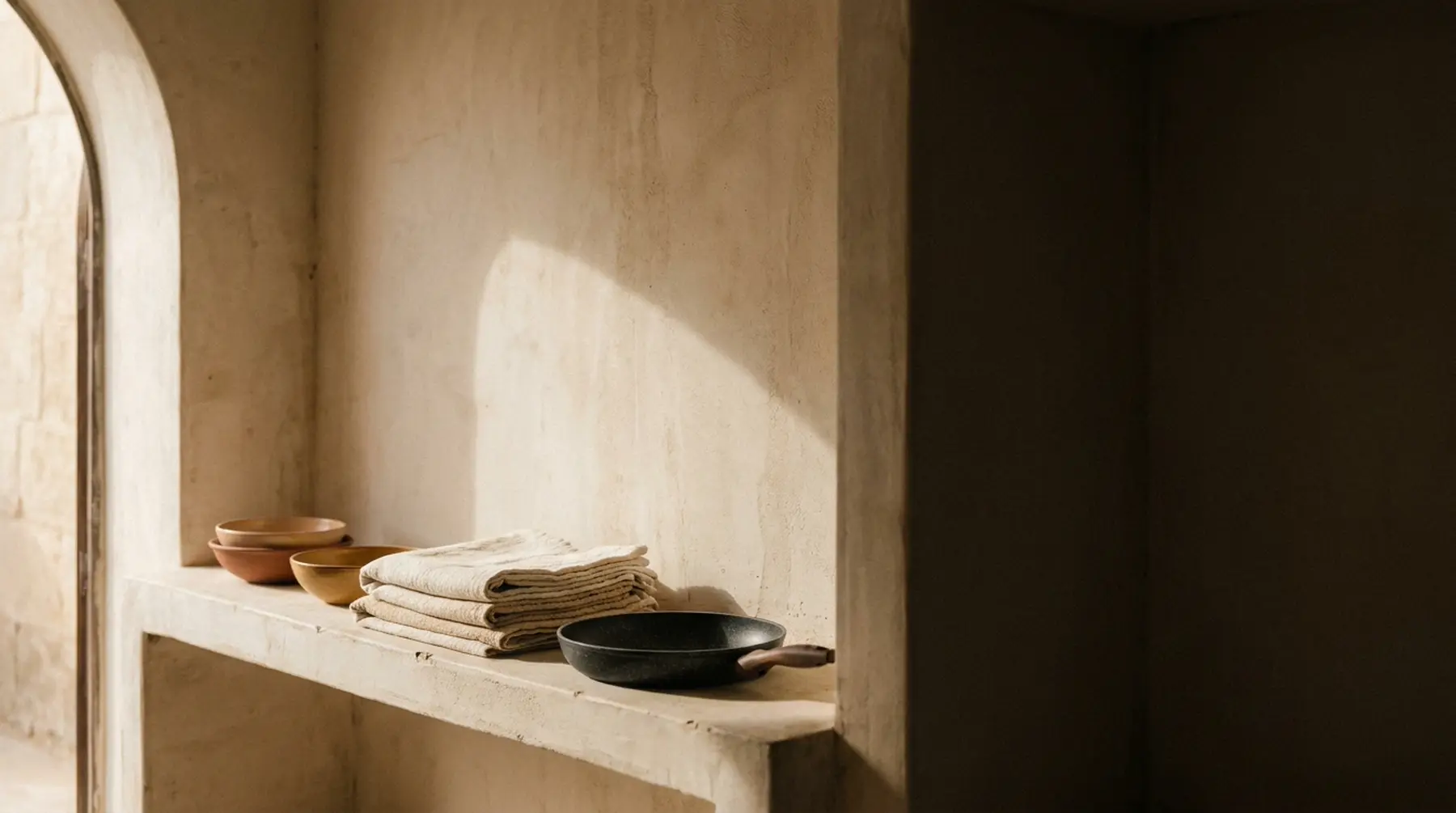 Stoneware bowls, folded cotton and cookware arranged on a plaster shelf